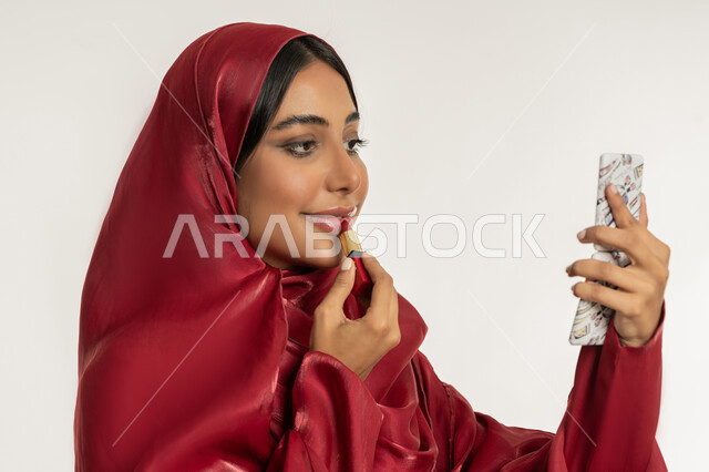العناية بالجمال والمظهر الخارجي, الترويج لمنتجات و مساحيق التجميل, استعمال المستحضرات التجميلية, مفهوم التزين والتجمل, بورتريه مقرب لامرأة عربية خليجية إماراتية محجبة ترتدي العباءة الحمراء تضع أحمر شفاه, شابة سعودية تقوم بوضع المكياج, خلفية بيضاء