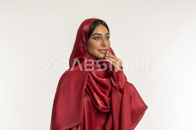 Promotion of cosmetics and products, use of cosmetics, concept of beautification and beautification, beauty care and appearance, close-up portrait of a veiled Emirati Gulf Arab woman wearing a red abaya and applying lipstick, a young Saudi woman applying makeup, white background