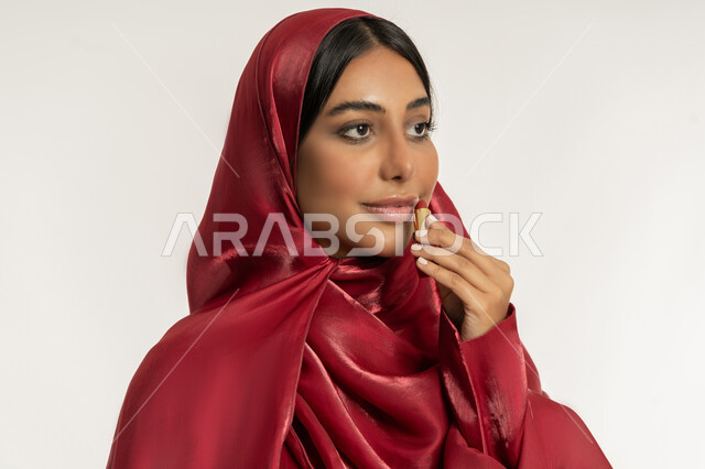العناية بالجمال والمظهر الخارجي, الترويج لمنتجات و مساحيق التجميل, استعمال المستحضرات التجميلية, مفهوم التزين والتجمل, بورتريه مقرب لامرأة عربية خليجية إماراتية محجبة ترتدي العباءة الحمراء تضع أحمر شفاه, شابة سعودية تقوم بوضع المكياج, خلفية بيضاء