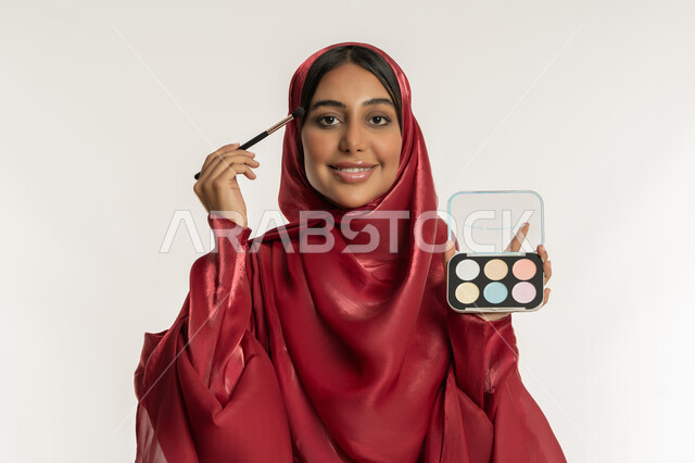 الاهتمام بالمظهر الخارجي, استخدام المستحضرات التجميلية, الترويج لمنتجات و مساحيق التجميل, مفهوم العناية بالجمال والبشرة, بورتريه مقرب لامرأة عربية خليجية إماراتية محجبة ترتدي العباءة الحمراء تستعمل فرشاة ظل العيون, شابة سعودية تقوم بوضع المكياج, خلفية بيضاء