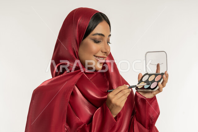 الترويج لمنتجات و مساحيق التجميل, استخدام المستحضرات التجميلية, الاهتمام بالمظهر الخارجي, مفهوم العناية بالجمال والبشرة, بورتريه مقرب لامرأة عربية خليجية إماراتية محجبة ترتدي العباءة الحمراء تستعمل فرشاة ظل العيون, شابة سعودية تقوم بوضع المكياج, خلفية بيضاء