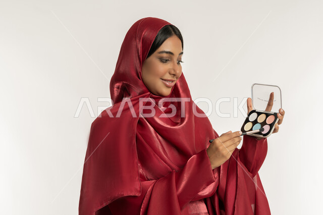 مفهوم العناية بالجمال والبشرة, الترويج لمنتجات و مساحيق التجميل, استخدام المستحضرات التجميلية, الاهتمام بالمظهر الخارجي, بورتريه مقرب لامرأة عربية خليجية إماراتية محجبة ترتدي العباءة الحمراء تستعمل فرشاة ظل العيون, شابة سعودية تقوم بوضع المكياج, خلفية بيضاء