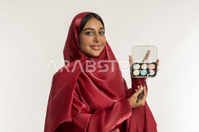 الاهتمام بالمظهر الخارجي, استخدام المستحضرات التجميلية, الترويج لمنتجات و مساحيق التجميل, مفهوم العناية بالجمال والبشرة, بورتريه مقرب لامرأة عربية خليجية إماراتية محجبة ترتدي العباءة الحمراء تستعمل فرشاة ظل العيون, شابة سعودية تقوم بوضع المكياج, خلفية بيضاء