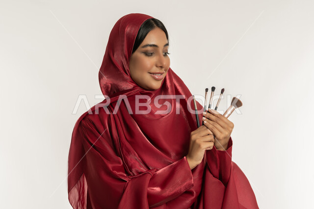 الترويج لمنتجات و مساحيق التجميل, استخدام المستحضرات التجميلية, مفهوم العناية بالجمال والبشرة, بورتريه مقرب لامرأة عربية خليجية إماراتية محجبة ترتدي العباءة الحمراء تمسك بيدها الفراشي وتنظر إليهم, شابة سعودية تستعمل أدوات المكياج, خلفية بيضاء
