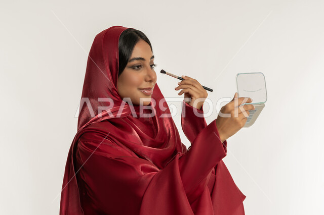 مفهوم العناية بالجمال والبشرة, الترويج لمنتجات و مساحيق التجميل, استخدام المستحضرات التجميلية, بورتريه مقرب من الجانب لامرأة عربية خليجية إماراتية محجبة ترتدي العباءة الحمراء تستعمل فرشاة ظل العيون, شابة سعودية تقوم بوضع المكياج, خلفية بيضاء
