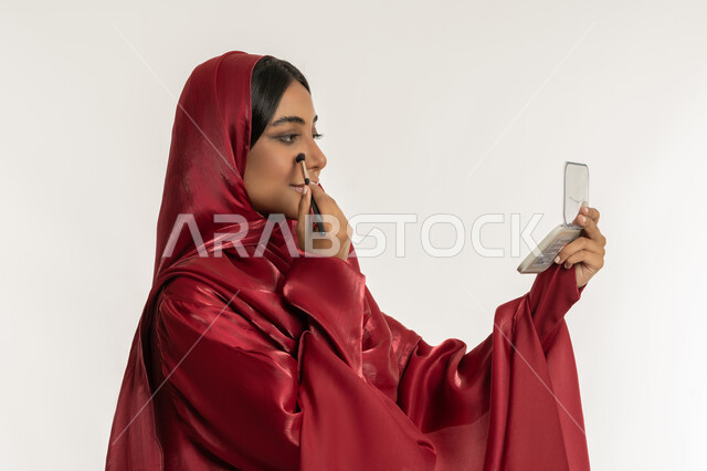 مفهوم التزين والتجمل, الاهتمام بالمظهر الخارجي, التسويق لمستحضرات ومنتجات التجميل, استخدام المساحيق التجميلية, بورتريه مقرب من الجانب لامرأة عربية خليجية إماراتية محجبة ترتدي العباءة الحمراء تستعمل فرشاة أحمر الخدود, شابة سعودية تضع المكياج, خلفية بيضاء