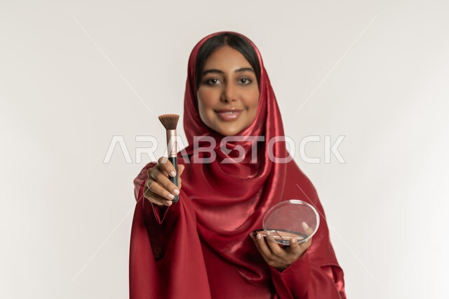 الاهتمام والعناية بالمظهر الخارجي, مفهوم التزين والتجمل, التسويق لمستحضرات ومنتجات التجميل, استخدام المساحيق التجميلية, بورتريه مقرب لامرأة عربية خليجية إماراتية محجبة ترتدي العباءة الحمراء تستعمل فرشاة أحمر الخدود, شابة سعودية تضع المكياج, خلفية بيضاء