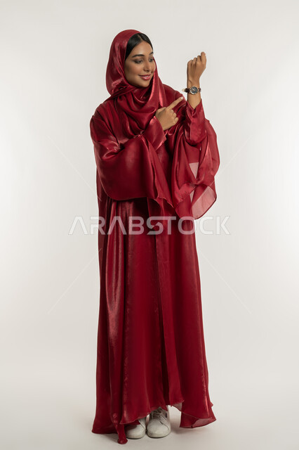 التذكير بموعد محدد, مفهوم الاهتمام بالأناقة والموضة, بورتريه لامرأة عربية خليجية إماراتية مبتسمة محجبة ترتدي العباءة الحمراء تضع ساعة يد وتشير إليها بإصبع السبابة, شابة سعودية تنظر إلى يدها, الجسم بالطول الكامل, خلفية بيضاء