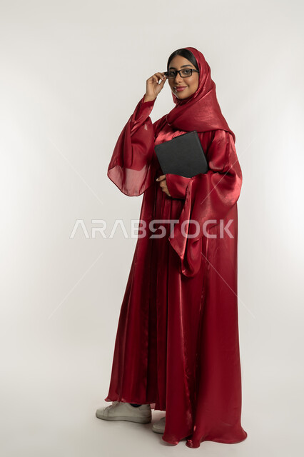 Integrating modern technologies and technology into practical life, portrait of a veiled Emirati Gulf Arab woman wearing a red abaya, wearing medical glasses and holding a tablet in her hand, a young Saudi woman looking at the camera with gestures of happiness and pleasure, full body, white background