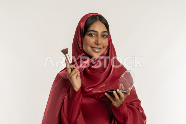 Use of cosmetics, concept of beautification and beautification, interest and care for external appearance, marketing of cosmetics and products, close-up portrait of a veiled Arab Gulf Emirati woman wearing a red abaya using a blush brush, a young Saudi woman applying makeup, white background