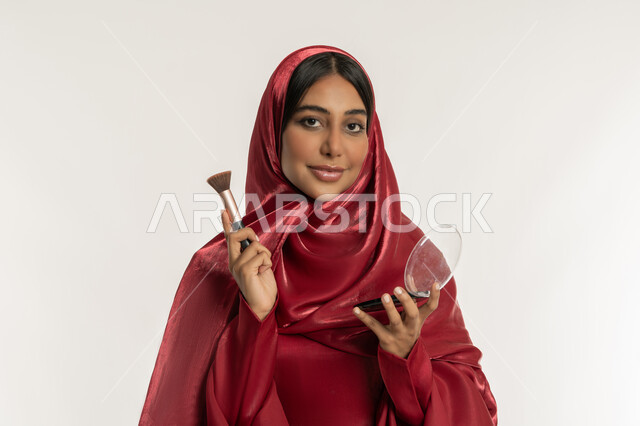 Marketing of cosmetics and products, use of cosmetics, care and attention to one's appearance, concept of beautification and beautification, close-up portrait of a veiled Emirati Gulf Arab woman wearing a red abaya using a blush brush, a young Saudi woman applying makeup, white background