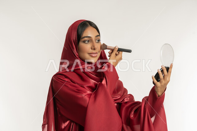 مفهوم التزين والتجمل, الاهتمام والعناية بالمظهر الخارجي, التسويق لمستحضرات ومنتجات التجميل, استخدام المساحيق التجميلية, بورتريه مقرب لامرأة عربية خليجية إماراتية محجبة ترتدي العباءة الحمراء تستعمل فرشاة أحمر الخدود, شابة سعودية تضع المكياج, خلفية بيضاء