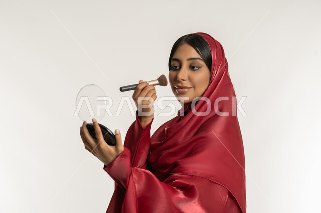 استخدام المساحيق التجميلية, التسويق لمستحضرات التجميل, الاهتمام والعناية بالمظهر الخارجي, مفهوم التزين والتجمل, بورتريه مقرب من الجانب لامرأة عربية خليجية إماراتية محجبة ترتدي العباءة الحمراء تستعمل فرشاة أحمر الخدود, شابة سعودية تضع المكياج, خلفية بيضاء