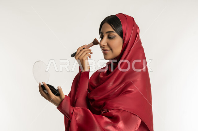 Marketing of cosmetics, use of cosmetics, care and attention to one's appearance, concept of beautification and beautification, close-up portrait from the side of a veiled Emirati Gulf Arab woman wearing a red abaya using a blush brush, a young Saudi woman applying makeup, white background