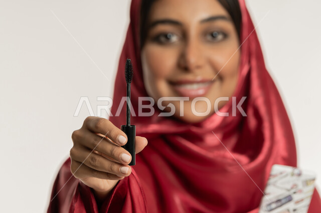 مفهوم التزين والتجمل, الشغف بوضع المكياج, التسويق لمنتجات ومستحضرات التجميل, بورتريه مقرب لامرأة عربية خليجية إماراتية محجبة ترتدي العباءة الحمراء تمسك بيدها فرشاة المسكرة, شابة سعودية تنظر إلى الكاميرا بإيماءات السعادة والسرور, خلفية بيضاء