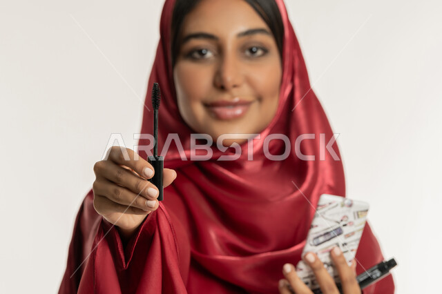 Passion for applying makeup, concept of beautification and beautification, marketing of cosmetics and products, close-up portrait of a veiled Emirati Gulf Arab woman wearing a red abaya holding a mascara brush in her hand, a young Saudi woman looking at the camera with gestures of happiness and pleasure, white background