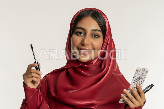 التسويق لمنتجات ومستحضرات التجميل, الشغف بوضع المكياج, مفهوم التزين والتجمل, بورتريه مقرب لامرأة عربية خليجية إماراتية محجبة ترتدي العباءة الحمراء تمسك بيدها فرشاة المسكرة, شابة سعودية تنظر إلى الكاميرا بإيماءات السعادة والسرور, خلفية بيضاء