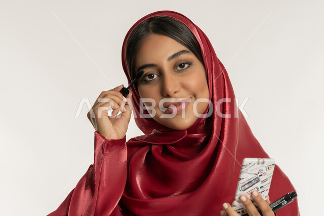 Concept of beauty care and appearance, passion for applying makeup, promotion of cosmetics and products, close-up portrait of a veiled Emirati Gulf Arab woman wearing a red abaya applying mascara, a young Saudi woman looking at the camera with gestures of happiness and pleasure, white background