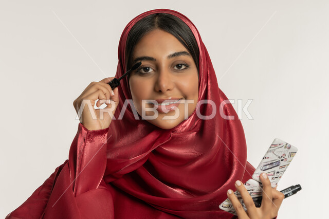 الشغف بوضع المكياج, مفهوم العناية بالجمال والمظهر الخارجي, الترويج لمنتجات ومستحضرات التجميل, بورتريه مقرب لامرأة عربية خليجية إماراتية محجبة ترتدي العباءة الحمراء تقوم باستخدام المسكرة, شابة سعودية تنظر إلى الكاميرا بإيماءات السعادة والسرور, خلفية بيضاء