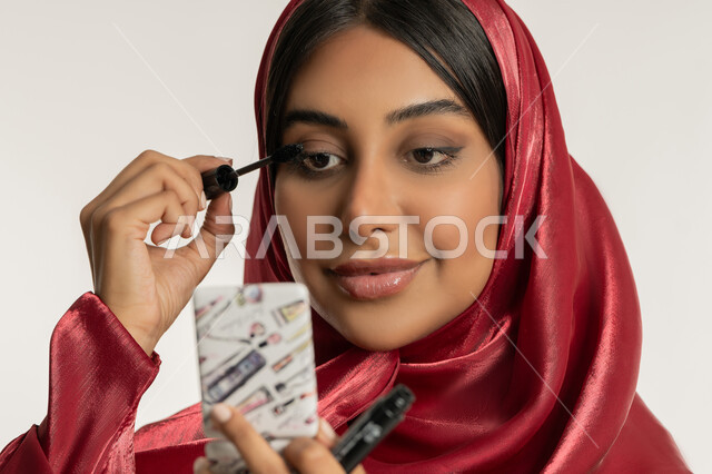 مفهوم التزين والتجمل, الاهتمام بالجمال والمظهر الخارجي, الترويج لمنتجات ومستحضرات التجميل, بورتريه مقرب لامرأة عربية خليجية إماراتية محجبة ترتدي العباءة الحمراء تقوم باستخدام المسكرة, شابة سعودية تضع المكياج, خلفية بيضاء