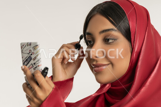 الاهتمام بالجمال والمظهر الخارجي, مفهوم التزين والتجمل, الترويج لمنتجات ومستحضرات التجميل, بورتريه مقرب لامرأة عربية خليجية إماراتية محجبة ترتدي العباءة الحمراء تقوم باستخدام المسكرة, شابة سعودية تضع المكياج, خلفية بيضاء