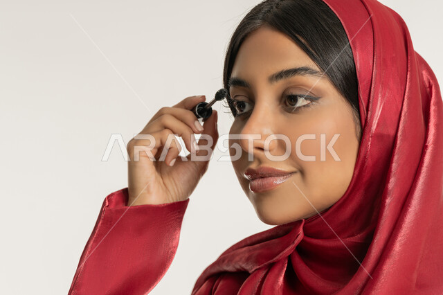 الترويج لمنتجات ومستحضرات التجميل, الاهتمام بالجمال والمظهر الخارجي, مفهوم التزين والتجمل, بورتريه مقرب لامرأة عربية خليجية إماراتية محجبة ترتدي العباءة الحمراء تقوم باستخدام المسكرة, شابة سعودية تضع المكياج, خلفية بيضاء