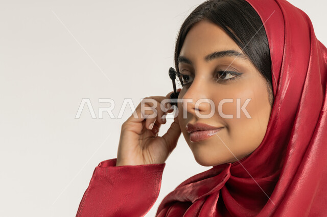 مفهوم التزين والتجمل, الاهتمام بالجمال والمظهر الخارجي, الترويج لمنتجات ومستحضرات التجميل, بورتريه مقرب لامرأة عربية خليجية إماراتية محجبة ترتدي العباءة الحمراء تقوم باستخدام المسكرة, شابة سعودية تضع المكياج, خلفية بيضاء