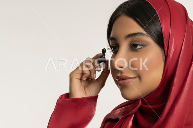 Caring about beauty and appearance, concept of adornment and beautification, promotion of cosmetics and products, close-up portrait of a veiled Emirati Gulf Arab woman wearing a red abaya using mascara, young Saudi woman applying makeup, white background