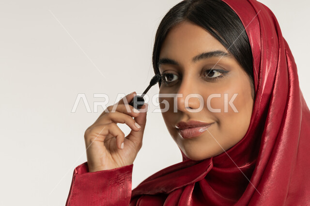 الترويج لمنتجات ومستحضرات التجميل, الاهتمام بالجمال والمظهر الخارجي, مفهوم التزين والتجمل, بورتريه مقرب لامرأة عربية خليجية إماراتية محجبة ترتدي العباءة الحمراء تقوم باستخدام المسكرة, شابة سعودية تضع المكياج, خلفية بيضاء