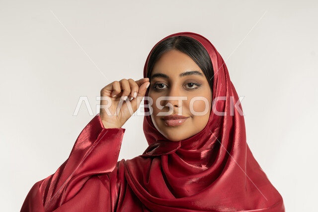 التسويق لمنتجات ومستحضرات التجميل, الاهتمام بالجمال والمظهر الخارجي, مفهوم التزين والتجمل, بورتريه مقرب لامرأة عربية خليجية إمارتية محجبة ترتدي العباءة الحمراء تقوم بوضع الرموش الاصطناعية, شابة سعودية تضع المكياج, خلفية بيضاء