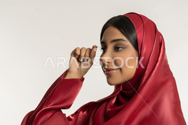 مفهوم التزين والتجمل, التسويق لمنتجات ومستحضرات التجميل, الاهتمام بالجمال والمظهر الخارجي, بورتريه مقرب لامرأة عربية خليجية إمارتية محجبة ترتدي العباءة الحمراء تقوم بوضع الرموش الاصطناعية, شابة سعودية تضع المكياج, خلفية بيضاء