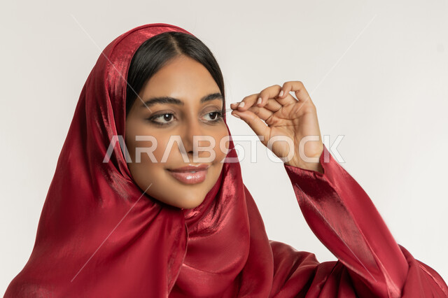 الاهتمام بالجمال والمظهر الخارجي, التسويق لمنتجات ومستحضرات التجميل, مفهوم التزين والتجمل, بورتريه مقرب لامرأة عربية خليجية إمارتية محجبة ترتدي العباءة الحمراء تقوم بوضع الرموش الاصطناعية, شابة سعودية تضع المكياج, خلفية بيضاء