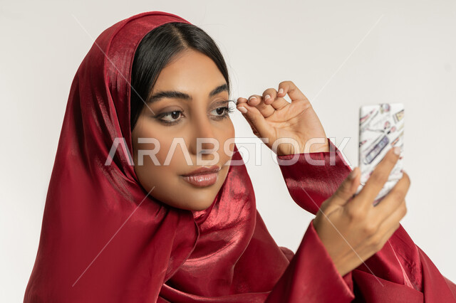 التسويق لمنتجات ومستحضرات التجميل, الاهتمام بالجمال والمظهر الخارجي, مفهوم التزين والتجمل, بورتريه مقرب لامرأة عربية خليجية إمارتية محجبة ترتدي العباءة الحمراء تقوم بوضع الرموش الاصطناعية, شابة سعودية تضع المكياج, خلفية بيضاء
