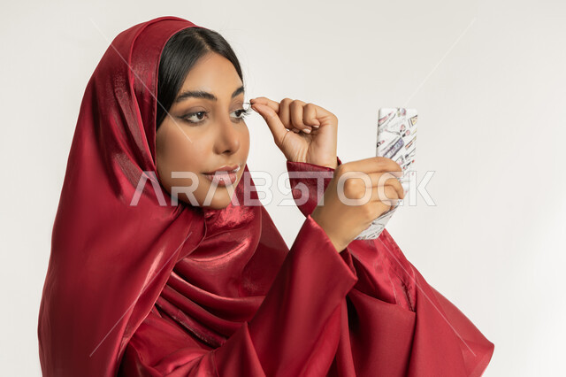 مفهوم التزين والتجمل, التسويق لمنتجات ومستحضرات التجميل, الاهتمام بالجمال والمظهر الخارجي, بورتريه مقرب لامرأة عربية خليجية إمارتية محجبة ترتدي العباءة الحمراء تقوم بوضع الرموش الاصطناعية, شابة سعودية تضع المكياج, خلفية بيضاء