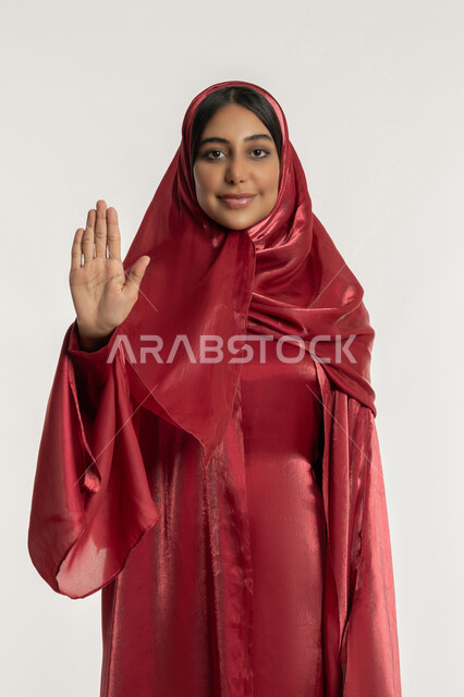 طلب التوقف والامتناع, تعابير الرفض وعدم الموافقة والقبول, بورتريه لامرأة عربية خليجية إماراتية ترتدي العباءة الحمراء ترفع يدها للأعلى, شابة سعودية تنظر إلى الكاميرا بإيماءات السعادة والسرور, خلفية بيضاء