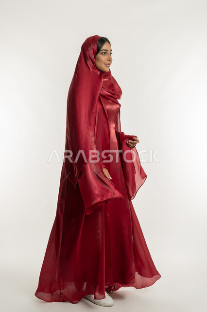 Concept of caring about elegance and appearance, modern and elegant veiled fashion, side portrait of an Arab Gulf Emirati woman wearing a red abaya, expressions of walking and going, a young Saudi woman looking at something with gestures of happiness and pleasure, full-length body, white background