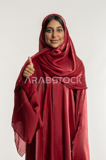 تعابير الرضا والموافقة, مفهوم القبول والإعجاب, بورتريه لامرأة عربية خليجية إماراتية ترتدي العباءة الحمراء تشير بإصبع الإبهام للأعلى, شابة سعودية تنظر إلى الكاميرا بإيماءات السعادة والسرور, خلفية بيضاء