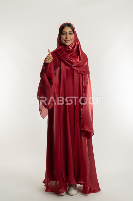 مفهوم القبول والإعجاب, تعابير الرضا والموافقة, بورتريه لامرأة عربية خليجية إماراتية ترتدي العباءة الحمراء تشير بإصبع الإبهام للأعلى, شابة سعودية تنظر إلى الكاميرا بإيماءات السعادة والسرور, الجسم بالطول الكامل, خلفية بيضاء