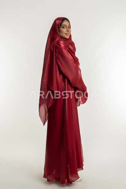 الاهتمام والعناية بالمظهر الخارجي, مفهوم الموضة والأناقة, بورتريه من الجانب لامرأة عربية خليجية إماراتية ترتدي العباءة الحمراء تقف بتعابير الحياء والنعومة, شابة سعودية تنظر إلى الكاميرا بإيماءات السعادة والسرور, الجسم بالطول الكامل, خلفية بيضاء