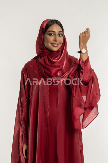 عرض ماركات ساعات وطنية فاخرة, مفهوم الاهتمام بالأناقة والموضة, بورتريه لامرأة عربية خليجية إماراتية مبتسمة محجبة ترتدي العباءة الحمراء وساعة يد ترفع يدها للأعلى, شابة سعودية تنظر إلى الكاميرا بإيماءات السعادة والسرور, خلفية بيضاء