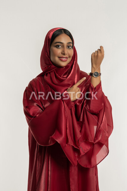مفهوم الاهتمام بالأناقة والموضة, التذكير بموعد محدد, بورتريه لامرأة عربية خليجية إماراتية مبتسمة محجبة ترتدي العباءة الحمراء تضع ساعة يد وتشير إليها بتعابير التأكيد على الالتزام بالوقت, شابة سعودية تنظر إلى الكاميرا بإيماءات السعادة والسرور, خلفية بيضاء