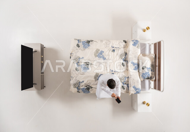 Benefiting from mobile applications and programs, using modern electronic devices, vertical overhead portrait of an Arab Gulf Omani man sitting on a comfortable bed holding a mobile phone in his hand, a Saudi young man browsing social networking sites, chatting and communicating with family and friends, white background