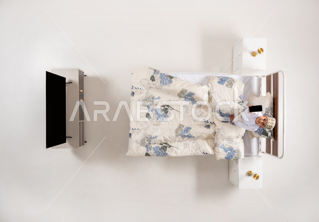 عرض شاشة سوداء فارغة، الاستفادة من تطبيقات وبرامج الموبايل، استخدام الأجهزة الالكترونية الحديثة، بورتريه علوي عامودي لولد عربي خليجي عماني يرتدي الدشداشة والكمة يجلس على سرير مريح يحمل بيده الهاتف المحمول، خلفية بيضاء