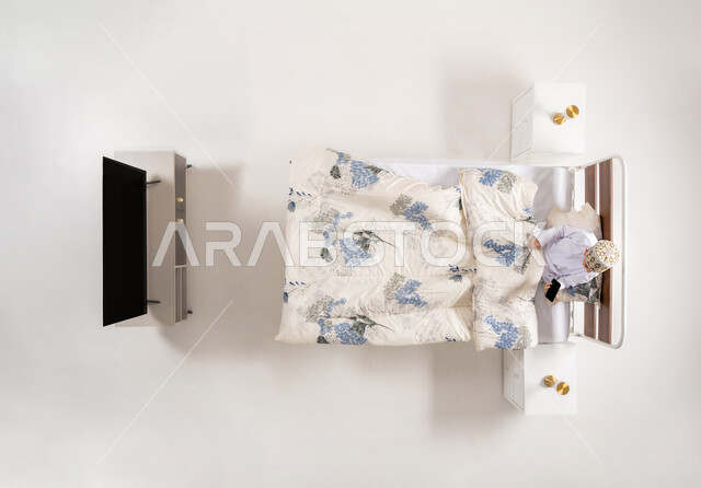 Using modern electronic devices, following entertainment programs via mobile, communicating with family and friends, vertical upper portrait of an Arab Gulf Omani boy wearing a dishdasha and a kimono sitting on a comfortable bed holding a mobile phone in his hand, white background