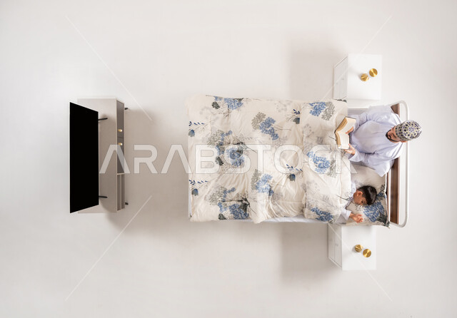 قضاء الوقت مع الابن، مفهوم التربيه القائمه على الحب والحنان، قراءه القصص المفيده قبل النوم، بورتريه علوي عامودي لاب عربي خليجي عماني يستلقي على سرير مريح برفقة ولده يقرأ الروايات المسليه، خلفية بيضاء