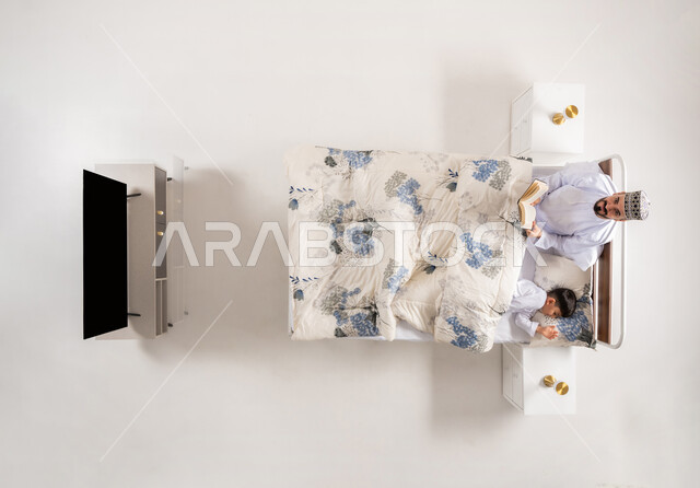 مفهوم التربيه القائمه على الحب والحنان، قضاء الوقت مع الابن، قراءه القصص المفيده قبل النوم، بورتريه علوي عامودي لاب عربي خليجي عماني يستلقي على سرير مريح برفقة ولده يقرأ الروايات المسليه، خلفية بيضاء