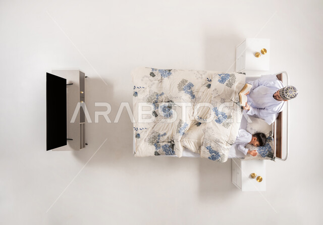 قراءه القصص المفيده قبل النوم، مفهوم التربيه القائمه على الحب والحنان، قضاء الوقت مع الابن، بورتريه علوي عامودي لاب عربي خليجي عماني يستلقي على سرير مريح برفقة ولده يقرأ الروايات المسليه، خلفية بيضاء