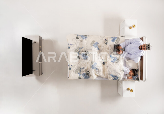 متابعة المشاريع والخطط عبر الانترنت، إنجاز مهام الأعمال عن بعد، استخدام الأجهزة التكنولوجية الحديثة والمتطورة، بورتريه علوي عامودي لرجل عربي خليجي عماني مستلقي على سرير مريح بجانب ابنه يعمل على جهاز الحاسوب المحمول، خلفيه بيضاء