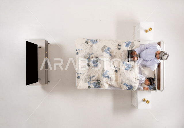 متابعة المشاريع والخطط عبر الانترنت، إنجاز مهام الأعمال عن بعد، استخدام الأجهزة التكنولوجية الحديثة والمتطورة، بورتريه علوي عامودي لرجل عربي خليجي عماني مستلقي على سرير مريح بجانب ابنه يعمل على جهاز الحاسوب المحمول، خلفيه بيضاء