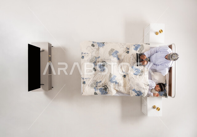 Completing business tasks remotely, using modern and advanced technological devices, vertical overhead portrait of an Arab Gulf Omani man lying on a comfortable bed next to his son working on a laptop, following up on projects and plans online, white background